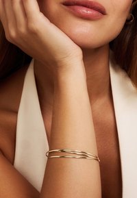 Bracelet jonc en ton or avec un design torsadé, comportant plusieurs bandes lisses et brillantes. Porté sur un poignet nu, mettant en valeur le teint de la peau.