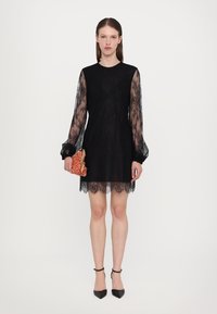 Rochie neagră cu mâneci lungi din dantelă și tiv din dantelă cu margine ondulată. Modelul ține în mână o poșetă texturată roșie și poartă pantofi negri cu vârful ascuțit.