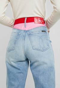 Person som bär ljusblå denimjeans med ett rosa midjeband och ett rött Calvin Klein-bälte, händerna vilande på höfterna, beige långärmad topp.