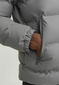 Grå pufferjakke med elastiske manchetter. Har en lynlåslomme i siden og en glat, polstret tekstur, der viser et moderne, funktionelt design.