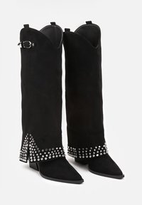 Bottes noires en velours côtelé montantes avec des pointes allongées, des clous en argent décoratifs autour de la base et une boucle sur le côté.