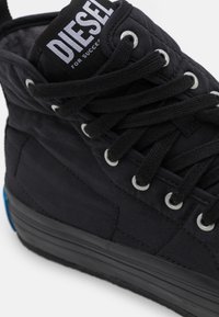 Czarne sneakersy za kostkę z tkaninową cholewką, teksturowanymi akcentami, białą etykietą z logo, czarnymi sznurówkami oraz grubą gumową podeszwą z niebieskim akcentem.