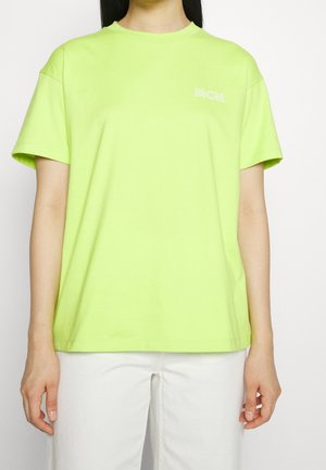 Osoba ubrana w jasną koszulkę z krótkim rękawem w kolorze limonkowej zieleni z logo "MCM" na piersi, zestawioną z białymi spodniami na białym tle.