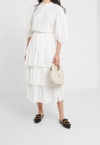 Gonna bianca a strati con pieghe, abbinata a una blusa bianca testurizzata. Scarpe nere slip-on con dettagli dorati. Borsa beige chiaro con manico circolare.