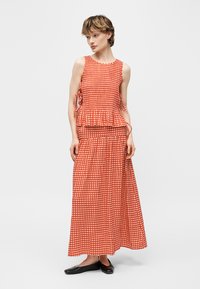 Vrouw die een mouwloze rood-wit geruite peplumtop draagt en een bijpassende lange rok met zwarte platte schoenen, staand tegen een witte achtergrond.