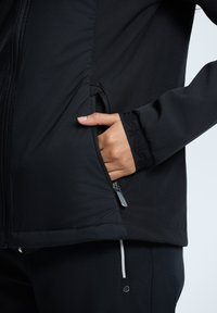 Veste noire à fermeture éclair avec une surface texturée, poche zippée et détail brodé aux poignets, associée à un pantalon noir assorti.