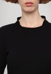 Crni pleteni top s teksturom, visokog okruglog izreza i raglan rukavima. Materijal ima suptilan rebrasti uzorak. Bez vidljivih kopčanja.