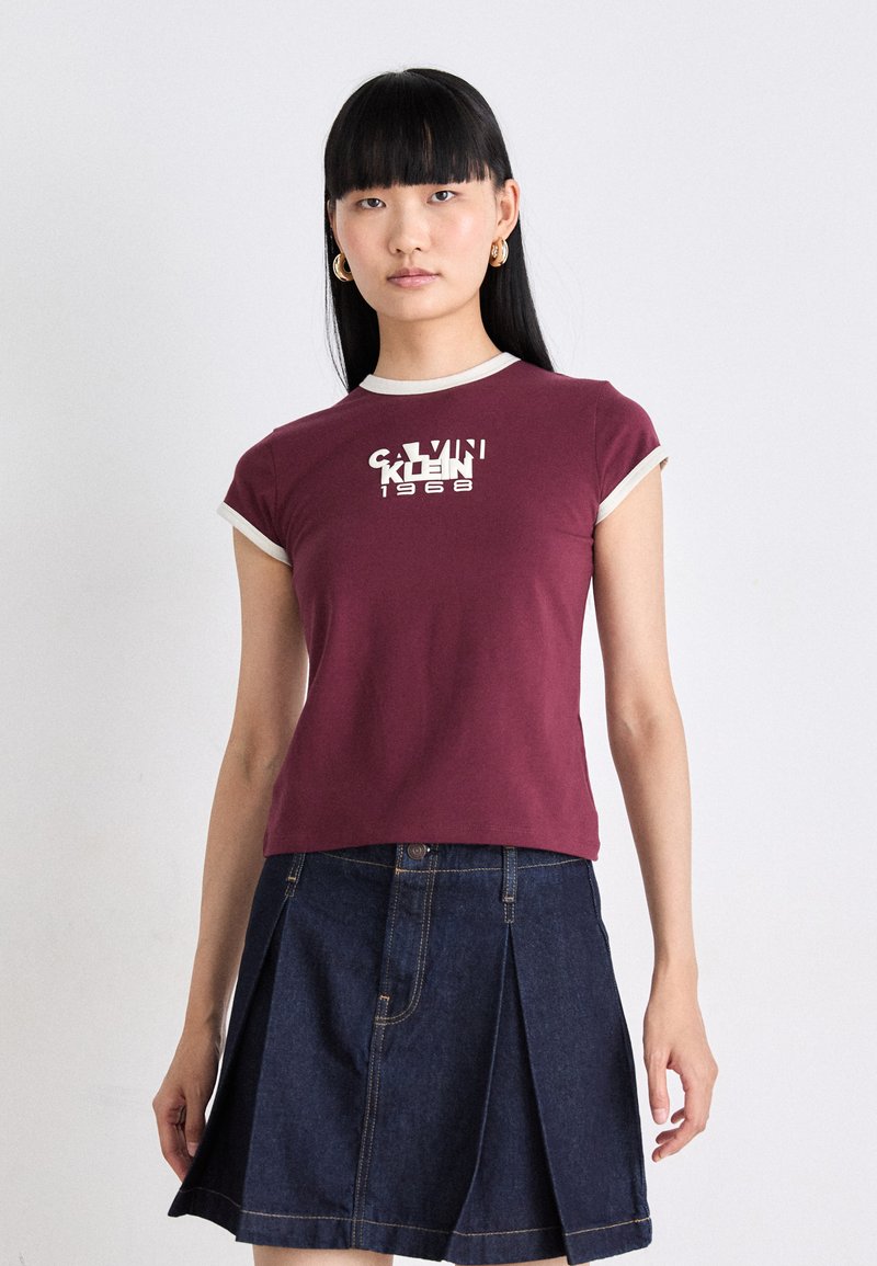 Maroon short-sleeve t-shirt with white trim and graphic logo. Paired with a dark denim skirt featuring a pleated design and front pockets.