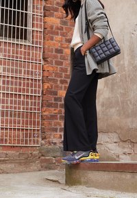 Schwarze weit geschnittene Hosen kombiniert mit einem beigen Pullover, einem karierten Blazer und bunten Sportschuhen. Eine schwarze gesteppte Tasche rundet das Outfit ab.