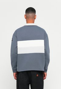 Mand med kort hår iført en blå og hvid farveblok sweatshirt, stående med ryggen til, parret med sorte bukser med en rød logo patch.