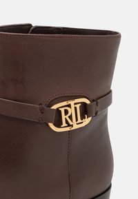 Gros plan sur une botte en cuir brun foncé avec une boucle logo "RL" dorée sur la sangle latérale, sur un fond uni.