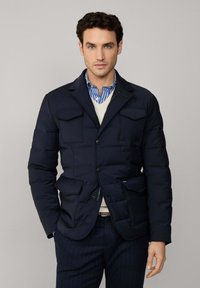 Marineblauwe gewatteerde jas met vier voorzakken, een geknipte kraag en knoopsluiting. Draagbaar over een lichte trui en een gestreept shirt, met een slim-fit ontwerp.