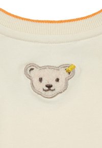 Cremfarbener Oberteil mit einem strukturierten Bärenkopf-Patch, umrandet von Grau, mit schwarzem Stick für Augen und Nase sowie einem orangen Halsabschluss.