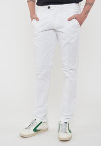 Pantaloni di cotone bianchi con taglio dritto, dotati di tasche frontali e chiusura con bottone. Abbinati a sneakers basse bianche e verdi.