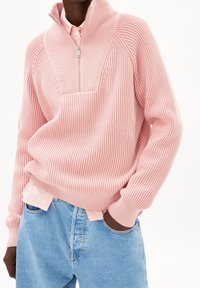 Pull en maille côtelée rose avec un col montant et une fermeture éclair mi-longue, superposé à une chemise rose clair. Associé à un jean en denim bleu clair.