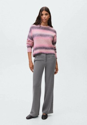 Eine Frau trägt einen pink- und lila gestreiften Pullover, graue weite Hosen mit seitlichen Nieten und schwarze, mit Nieten verzierte Slipper, während sie vor einem schlichten Hintergrund steht.