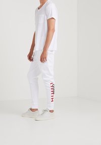Witte t-shirt met korte mouwen en een zak, gecombineerd met witte sweatpants met een rode tekstontwerp, en lichte sneakers. Schoon, minimalistische look.