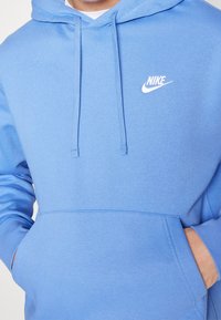 Ljusblå hoodie i mjukt tyg med en framficka och dragsnören. Har en vit Nike-logotyp på övre vänstra bröstet.