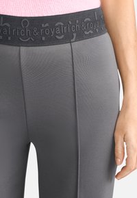 Primo piano di una persona che indossa pantaloni grigi con "rich & royal" stampato sulla cintura elastica nera e una maglietta rosa visibile sopra.