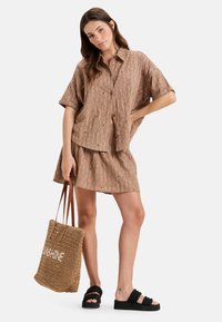 Bruine gestructureerde overhemd met korte mouwen, met knopen aan de voorkant, bijpassende rok, zwarte plateau-sandalen en een geweven tote bag met de tekst "SUNSHINE".