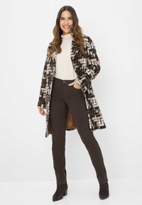 Cappotto lungo a righe in bianco e nero con una texture morbida, abbinato a pantaloni aderenti marroni scuri e stivaletti neri alla caviglia.