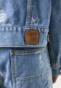 Veste en denim bleu avec un patch en cuir marron, des coutures visibles et des impressions graphiques subtiles sur le tissu.