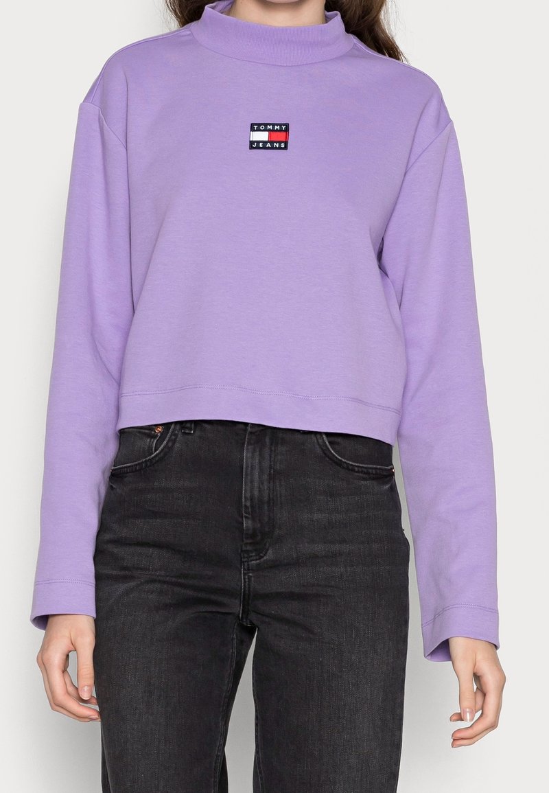 Femme portant un sweat court à manches longues lavande avec le logo "Tommy Jeans" et un jean noir taille haute sur un fond uni.