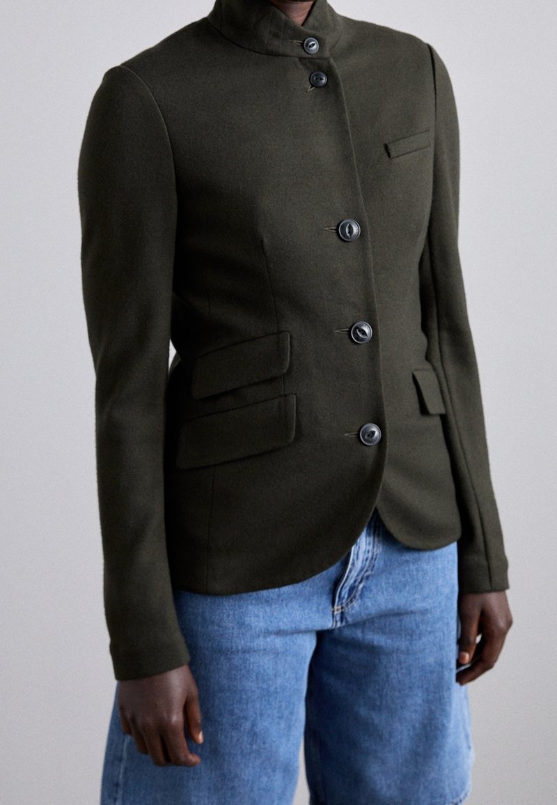 Personne portant une veste ajustée boutonnée vert foncé avec col montant et un jean bleu, debout devant un fond clair uni.