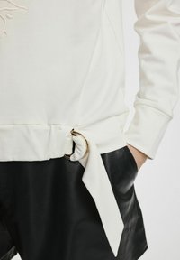 Biała bluza z węzłem i złotym akcentem w postaci pierścienia w talii, zestawiona z czarnymi szortami z ekoskóry. Widoczna miękka faktura materiału.