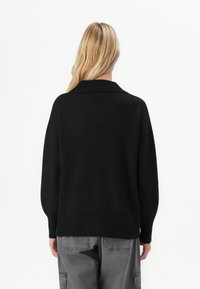 Mulher com cabelo loiro longo, vestindo um suéter de malha preto e calças cargo cinzas, de pé, de frente para um fundo branco simples.