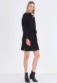 Zwarte jurk met lange mouwen, zijsplitsen en een tailleband. Glad materiaal en een hoge halslijn. Combinatie met zwarte enkellaarsjes.