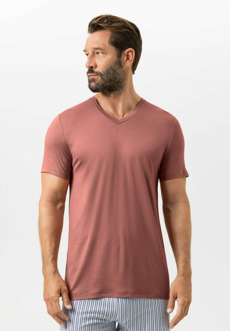 Uomo con barba che indossa una maglietta rosa polvere a scollo a V e pantaloni a righe, che guarda verso sinistra su uno sfondo chiaro uniforme.