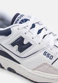 Zapatilla deportiva blanca con acentos en azul marino, que presenta una textura perforada, lengüeta acolchada y un destacado logo de 'N'. Número de modelo 550 visible.