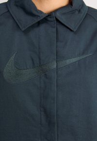 Ciemnoniebieska kurtka Nike z dużym, haftowanym czarnym logo swoosh z przodu. Posiada klasyczny kołnierz i gładką teksturę.