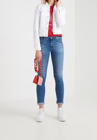 Hvid denimjakke, rød grafisk T-shirt, blå skinny jeans med opbrettede bukseben, hvide sneakers og en lille rød-og-hvid stribet håndtaske.