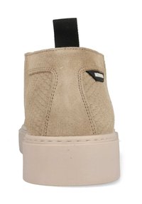 Tan suede schoen met gestructureerde zijkanten, dikke beige zool, zwarte stoffen trekstrip en klein zwart merkembleem op het hielgedeelte.
