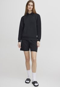 Svart hettegenser med lomme på forsiden, kombinert med svarte shorts. Modellen har på seg hvite sokker og svarte sandaler. Enkelt, uformelt design.
