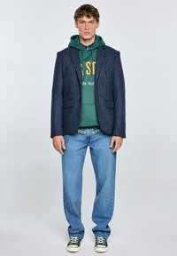 Mornarska jakna z teksturiranim zaključkom, zeleni kapucar z logotipom, ovratnik iz karirane srajce, svetlosinji jeans in zeleni športni copati.