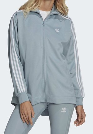Femme portant un survêtement Adidas bleu clair avec le détail des trois bandes blanches sur les manches et le pantalon, debout les mains détendues le long du corps.