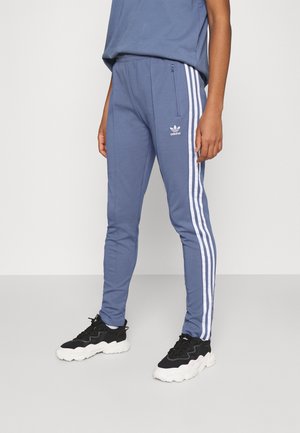 Osoba nosząca niebieskie spodnie dresowe Adidas z białymi paskami po bokach oraz czarno-białe trampki, stojąca na jednolitym tle.