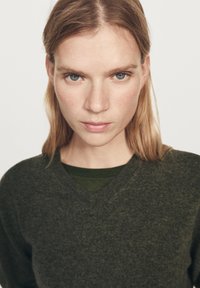 Jeune femme à la peau claire et aux cheveux blonds, portant un pull vert foncé, regardant droit devant avec une expression sérieuse, sur fond blanc uni.