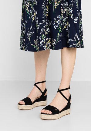 Kvinde iført sorte kilehæls-sandaler med ankelremme og en navyblå blomsterprint midi-nederdel, stående på et gråt gulv foran en hvid væg.