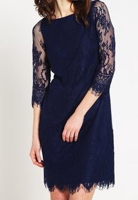 Marinblå spetsklänning med ärmar i trekvarts längd av genomskinlig spets, vågigt fåll och intrikata blommönster över ett slätt underlager.