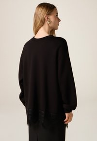 Sudadera negra con escote redondeado y mangas voluminosas. Cuenta con un borde de encaje en la parte inferior con patrones intrincados.