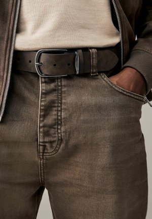 Jeans de mezclilla marrones con un acabado texturizado y un ligero desgaste, combinados con un cinturón de cuero oscuro que tiene una hebilla de metal y detalles de costura visibles.