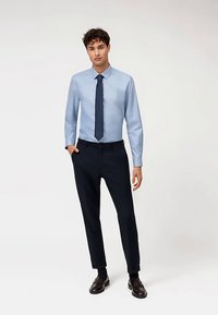 Hellblaues Hemd mit Knopfleiste und spitzem Kragen, kombiniert mit dunkelblauen, schlanken Hosen und braunen Mokassins. Sauberes Design, ohne sichtbare Muster.