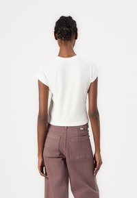 Rövid ujjú, rövid fazonú fehér póló, magasan szabott, bőszárú mályva színű nadrággal, mely két hátsó zsebbel rendelkezik.