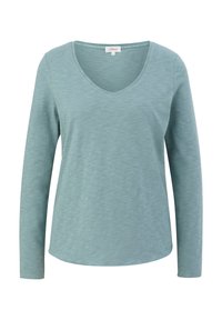 Hellblaues Langarmshirt mit V-Ausschnitt, aus strukturiertem Stoff und mit abgerundetem Saum. Das Etikett innen trägt den Aufdruck "LASST UNS NETT ZUEINANDER SEIN".