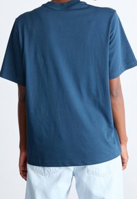 Osoba v jednoduchém tmavě modrém tričku s krátkým rukávem a světle modrých džínách, zachycena zezadu na jednobarevném bílém pozadí.