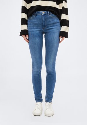 Person wearing slim-fit blue jeans, white sneakers, and a black and white striped long-sleeve sweater, standing against a plain background.